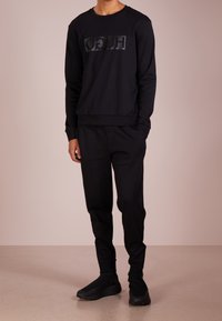 Schwarzer Sweatshirt mit einem glänzenden schwarzen "HUGO"-Logo, kombiniert mit schwarzen Jogginghosen und schwarzen Sneakern. Glatter Stoff und lockere Passform.