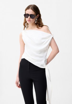 Kobieta z falującymi brązowymi włosami, nosząca czarne okulary przeciwsłoneczne, białą asymetryczną bluzkę bez rękawów oraz czarne dopasowane spodnie na białym tle.