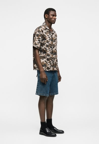 Άνδρας που στέκεται φορώντας καφέ κοντομάνικο πουκάμισο παραλλαγής με κουμπιά, μπλε τζιν σορτς, μαύρες κάλτσες και μαύρα δερμάτινα μοκασίνια.