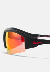 Occhiali da sole sportivi con montatura nera, accenti rossi e lenti arancioni. Presentano il marchio Nike e un design curvo e aerodinamico.