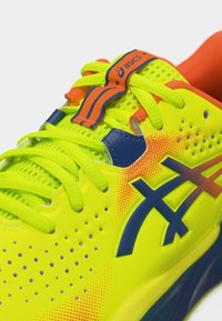Φωτεινό κίτρινο και πορτοκαλί αθλητικό παπούτσι ASICS με μπλε λογότυπο, κορδόνια νέον κίτρινου χρώματος και διάτρητο επάνω μέρος για αερισμό.
