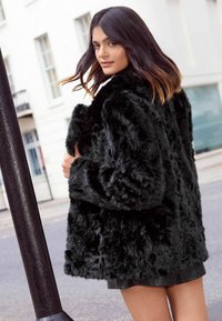 Melnas mākslīgās ādas mētelis ar teksturētu virsmu, platām piedurknēm un brīvu piegriezumu, kopā ar īsu melnu svārku.