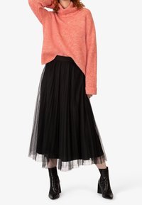 Maglione a collo alto in maglia corallo sovrapposto a una gonna maxi in tulle plissettato nera. Stivaletti neri con tacco completano l'outfit.