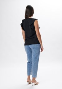 Schwarzes Oberteil mit Rüschensäumen, kombiniert mit hellblauen geraden Jeans. Das Modell hat lange Haare und trägt weiße High Heels.