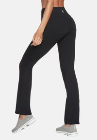 Schwarze Leggings mit einer taillierten Passform, ausgestattet mit Taschen auf der Rückseite und einem hohen Bund. Kombiniert mit grauen Slippern.