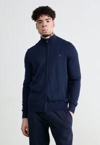 Suéter navy de cremallera con un alto cuello, puños y dobladillo acanalados, que presenta un pequeño logo en el lado izquierdo del pecho y una textura de punto suave.