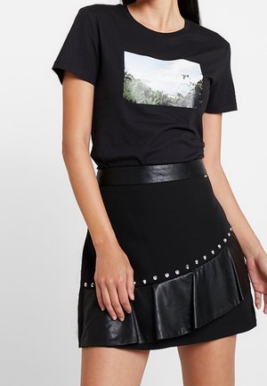 Frau trägt ein schwarzes T-Shirt mit kurzen Ärmeln und Naturgrafik sowie einen schwarzen asymmetrischen Rock mit Lederbesatz und silbernen Nieten.