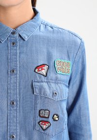 Camicia di jeans con chiusura a bottoni e tasca sul petto, decorata con toppe che includono due diamanti rossi, il testo "GOOD VIBES", un piccolo libro e un'ape.