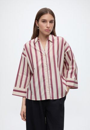 Femme aux longs cheveux bruns portant une chemise ample crème avec des rayures verticales grenat et un pantalon noir, debout devant un fond uni.