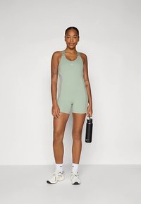 Zöld sportos egyrészes outfit versenyháttal és világosszürke díszítésekkel, fehér zoknival és fekete tornacipővel viselve, egy vizespalackot tartva.