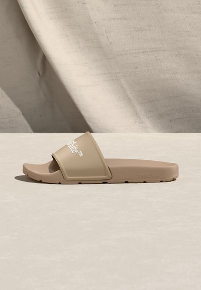 Μπεζ σανδάλι slide με φαρδιά λουρί και λευκό λογότυπο «Off-White», επίπεδη σόλα, τοποθετημένο σε φωτεινή επιφάνεια με υφασμάτινο φόντο.