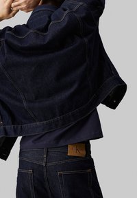 Mörkjeansjacka med avslappnad passform, med kontrasterande sömmar och en bred krage. Synlig brun läderlapp på jeansen.