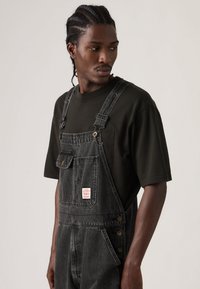 Νεαρός άνδρας με πλεγμένα μαλλιά φορώντας μαύρη τζιν φορμα συνοδευόμενη από μαύρο μπλουζάκι, κοιτάζει προς τα αριστερά του μπροστά σε ένα απλό φόντο.