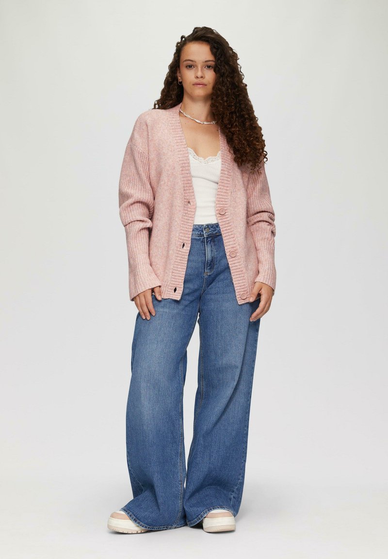 Różowy kardigan z prążkowanej dzianiny o luźnym kroju, zapinany na guziki, noszony na białej bluzce z koronkowym wykończeniem i w połączeniu z niebieskimi jeansami o szerokich nogawkach.