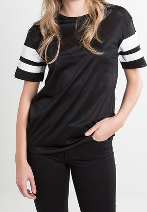 Mujer joven con una camiseta negra de manga corta con doble raya blanca en las mangas y pantalones negros, de pie con una mano en el bolsillo.