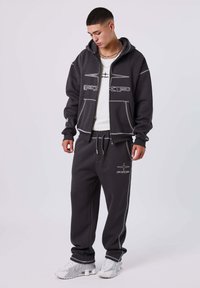 Joven con sudadera negra con cremallera y pantalones deportivos a juego, con zapatillas blancas, mirando hacia abajo contra un fondo liso claro.