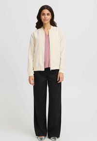 Light beige bomber jacket with a textured finish, zip closure, and ribbed cuffs, paired with a pink top and black wide-leg pants.