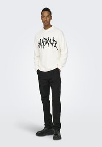 Maglione crema con grafica testuale nera "SHADOWS", abbinato a pantaloni neri e scarpe nere. Fit casual, maniche lunghe, orlo a costine.