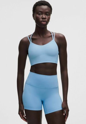 Mujer que lleva un top corto sin mangas color azul claro con doble tirante en los hombros y shorts ajustados de cintura alta a juego, de pie frente a un fondo liso.