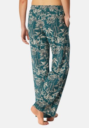 Persona descalza con pantalones color verde azulado con estampado botánico tropical blanco, vista desde atrás, de pie con una mano en el bolsillo.