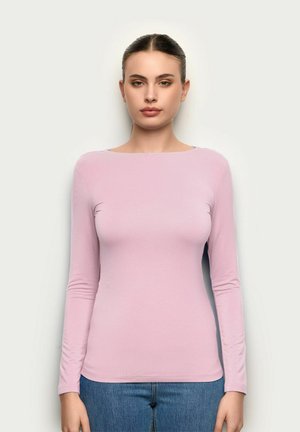 Kobieta o brązowych włosach związanych w gładki kok, ubrana w jasnoróżowy top z długim rękawem i niebieskie dżinsy, stojąca na tle jednolitego, jasnego tła.