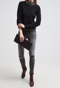 Czarny futrzany sweter z wysokim kołnierzem ozdobionym ćwiekami, parowany z szarymi przetarciami skinny jeans i ciemnobrązowymi botkami do kostek, trzymający czarną kopertówkę.