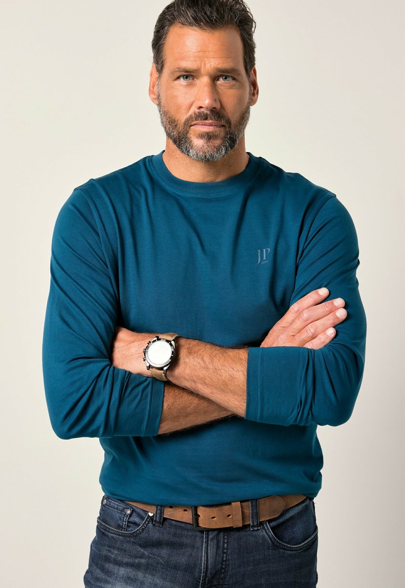 Teal langek mouwen shirt met een ronde hals en een klein logo. G gedragen met donkere jeans en een horloge. Heeft een gladde stof en aansluitende mouwen.