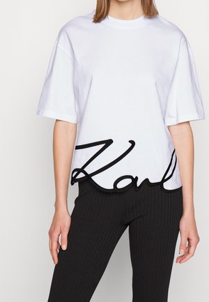 Woman wearing a white oversized T-shirt with black cursive text "Karl" across the waist, paired with black ribbed pants.