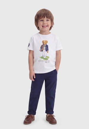 Băiat tânăr zâmbitor purtând un tricou alb cu un desen cu un ursuleț îmbrăcat, pantaloni bleumarin și pantofi maro cu șireturi.
