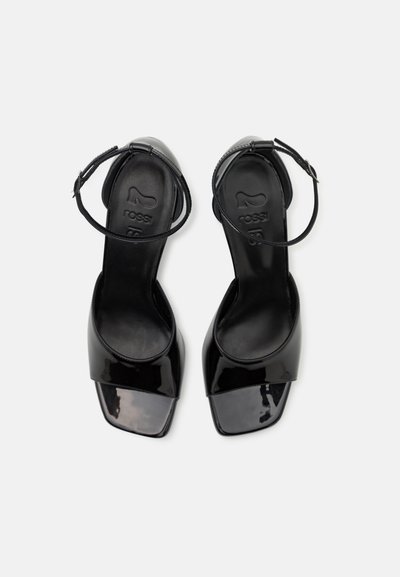 Black patent leather heels with a square toe design, ankle strap, and smooth texture, featuring a logo embossed on the insole.