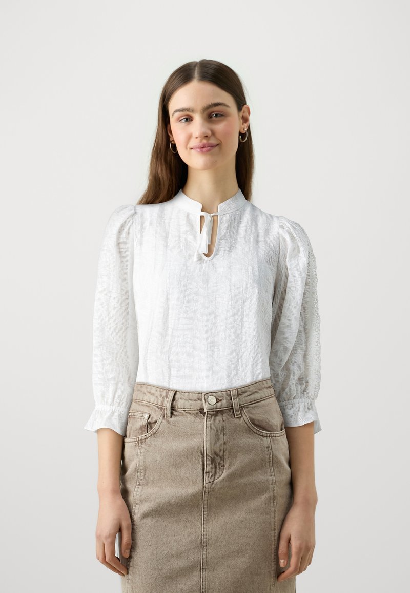 Vit texturerad blus med puffärmar och detalj i form av ett nyckelhål vid halsen, matchad med en taupe denimkjol.
