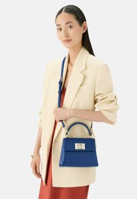 Blauwe leren handtas met een gestructureerde vorm, bovenhandgreep, afneembare schouderriem en zilveren hardwareaccenten. G gedragen bij een oversized crèmekleurige blazer.