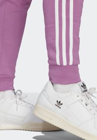 Lavendelfarbene Jogginghosen mit weißen Seitenstreifen, kombiniert mit weißen Ledersneakern mit beiger Sohle und schwarzen Logoakzenten.