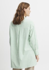 Camicia a righe verde e bianca con vestibilità rilassata, dettagli con polsini a bottoni e una cucitura sul retro. Tessuto di cotone morbido.