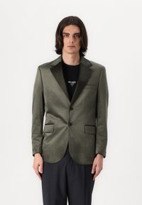 Olivgrüner Blazer mit satinierter Oberfläche, Ein-Knopf-Verschluss, Reverskragen und zwei vorderen Taschen. Über einem schwarzen T-Shirt getragen.