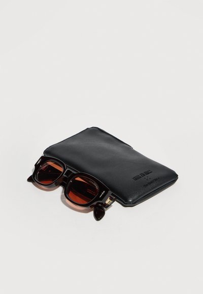 Μαύρα ορθογώνια γυαλιά ηλίου με σκούρους κόκκινους φακούς τοποθετημένα πάνω σε μαύρη δερμάτινη θήκη σε λευκό φόντο.