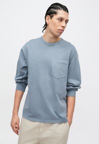 Μακρυμάνικο T-shirt σε ανοιχτό γαλάζιο χρώμα, κατασκευασμένο από μαλακό ύφασμα, με στρογγυλή λαιμόκοψη και τσέπη στο αριστερό μέρος του στήθους. Χαλαρή γραμμή και ριμπ μανσέτες.