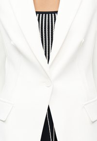 Λευκό προσαρμοσμένο σακάκι με κούμπωμα μονό κουμπί πάνω από μαύρο και λευκό πλεκτό τοπ με κάθετες ρίγες.