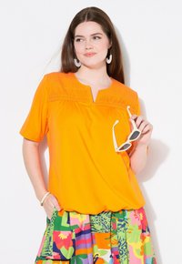 Oranžový top s krátkým rukávem a předními sklady, doplněný vícbarevnou květinovou sukní. Modelka drží bílé sluneční brýle.