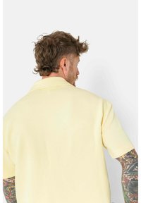 Polo de manga corta amarillo con una tela texturada y un collar clásico, mostrado desde la parte de atrás, revelando detalles de tatuajes coloridos en el brazo.