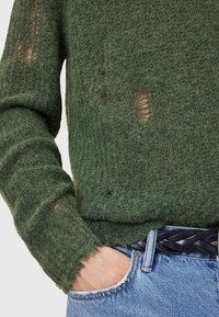 Zielony dzianinowy sweter z teksturowym wykończeniem, z dziurami i postrzępionymi brzegami. Połączony z niebieskimi dżinsami i plecionym czarnym paskiem.