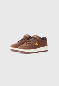 Adidași din piele maro cu talpă albă, având o curea cu velcro, șireturi maronii și un accent logo portocaliu pe lateral.