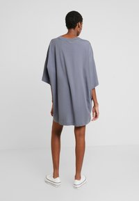 Kvinde iført en oversized grå T-shirt kjole med kortærmer og hvide platform sneakers, stående med ryggen til mod en ensfarvet lys baggrund.