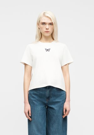 Junge Frau mit glattem, blondem Haar trägt ein weißes T-Shirt mit einem kleinen Schmetterlingsmotiv und blaue High-Waist-Jeans.