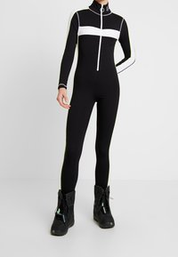 Svart helkroppsdräkt med vita horisontella ränder, hög krage och dragkedja framtill. Materialet verkar vara stretchigt. Bärs med svarta stövlar med snörning.