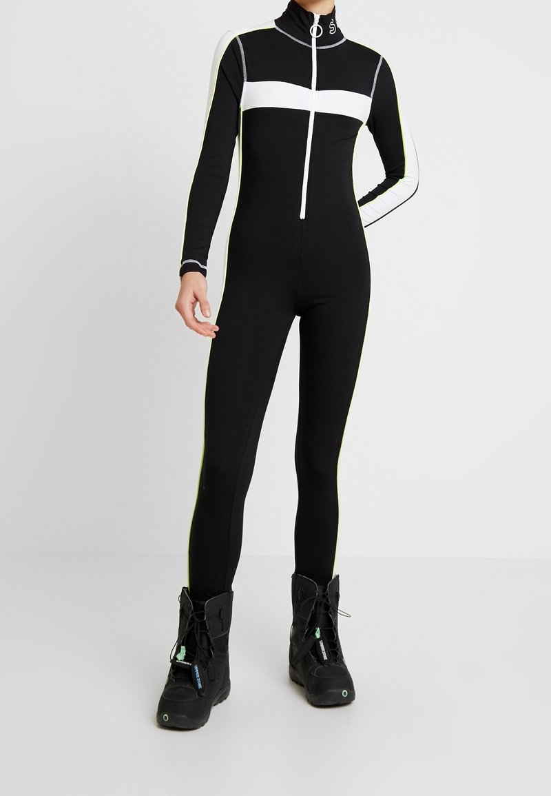 Svart helkroppsdräkt med vita horisontella ränder, hög krage och dragkedja framtill. Materialet verkar vara stretchigt. Bärs med svarta stövlar med snörning.