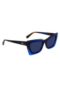 Ochelari de soare albaștri cu formă de pisică, ramă îndrăzneață albastră, lentile întunecate și accente de broască țestoasă multicolore pe brațe.