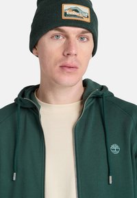 Felpa verde con zip e texture a coste, dotata di cappuccio e di un piccolo logo ricamato. Abbinata a una maglietta color crema e a un berretto verde scuro.