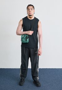 Top negru fără mâneci, pantaloni de trening negri cu dungi albe și adidași negri. Ținând o geantă mică de umăr verde cu fermoare.
