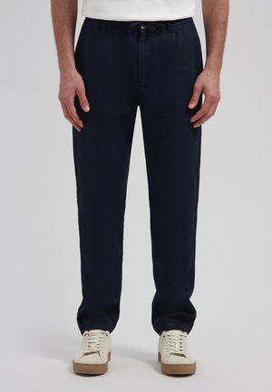 Hombre con pantalones azul marino de pierna recta y zapatillas blancas, de pie frente a un fondo liso.